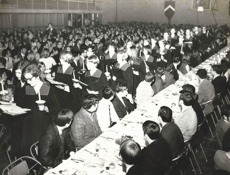 A large gathering of several hundred students, some sitting at long tables, others processing wearing robes and carrying candles.